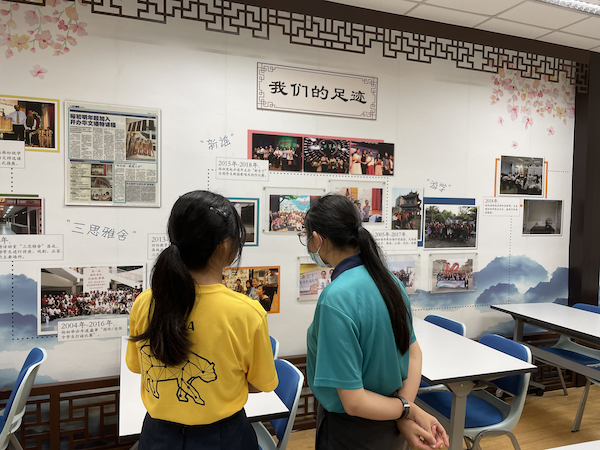 Two students stand looking at a Chinese-language display wall covered with photos and newspaper clippings.